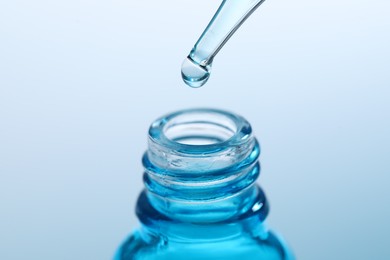 Dripping liquid from pipette into glass bottle on light blue background, closeup