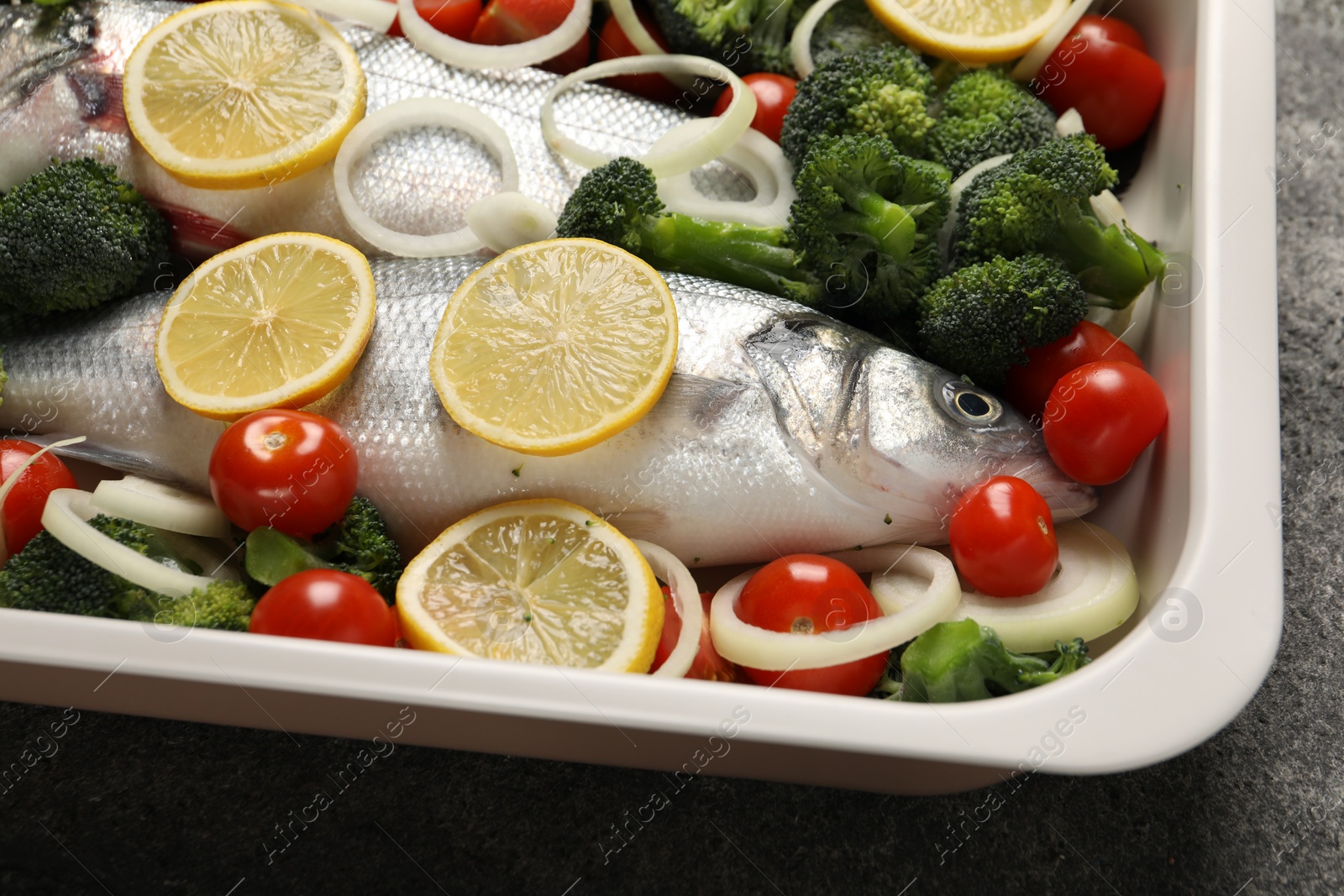 Photo of Raw fish with vegetables and lemon in baking dish on grey table