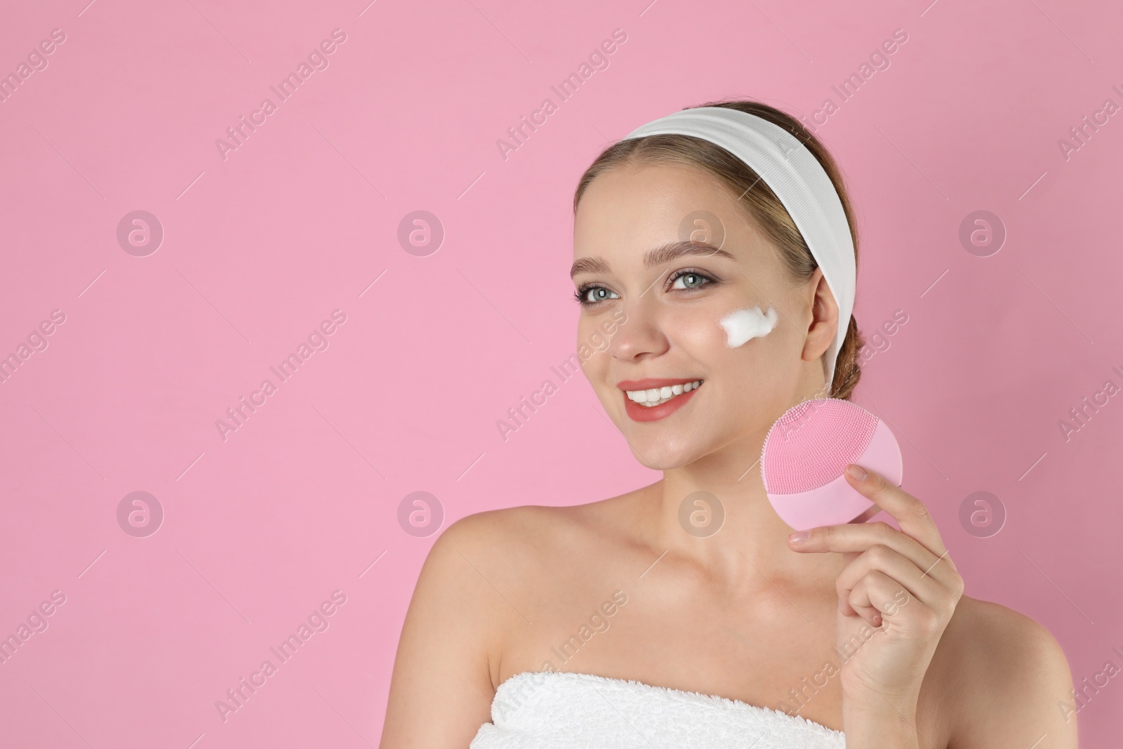 Photo of Young woman washing face with brush and cleansing foam on pink background, space for text. Cosmetic product