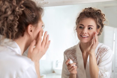 Beautiful woman applying moisturizing cream near mirror in bathroom