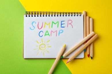 Notebook with written text SUMMER CAMP and different pencils on color background, top view