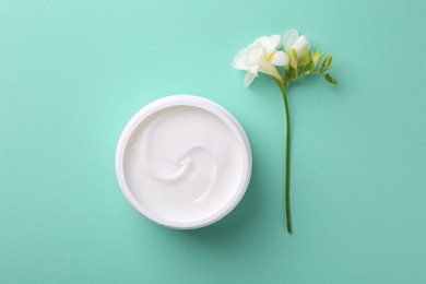 Moisturizing cream in open jar and freesia flower on turquoise background, flat lay. Body care product