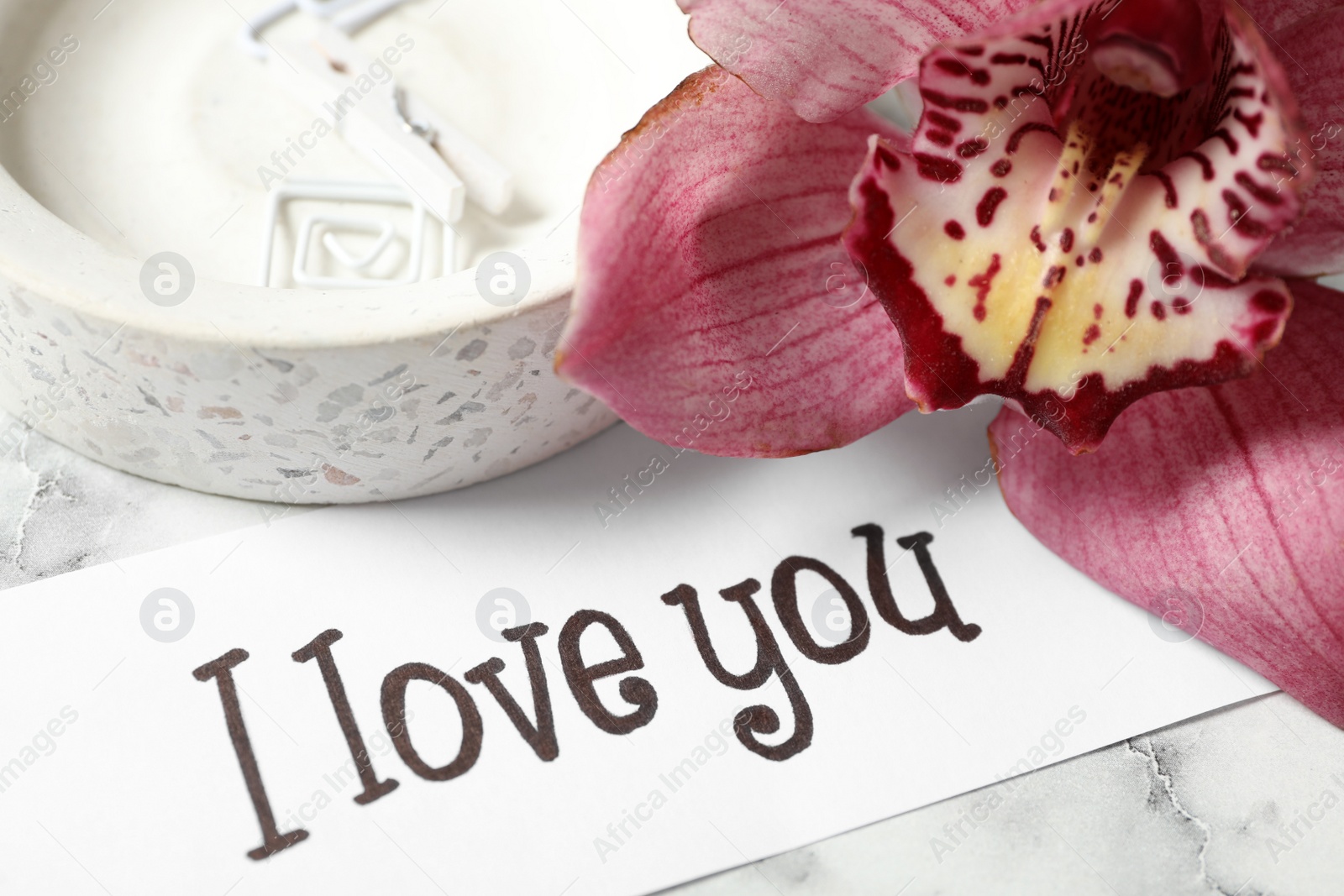Photo of Card with text I Love You, orchid flower and tray on white marble table, closeup
