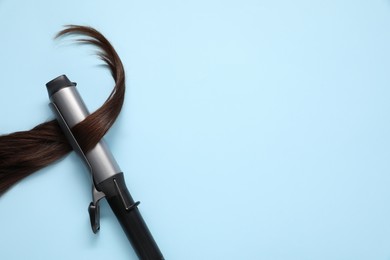 Photo of Curling iron with brown hair lock on light blue background, top view. Space for text