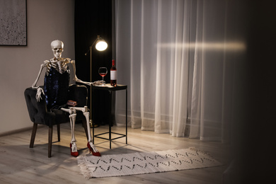 Skeleton in dress with wine sitting at table indoors