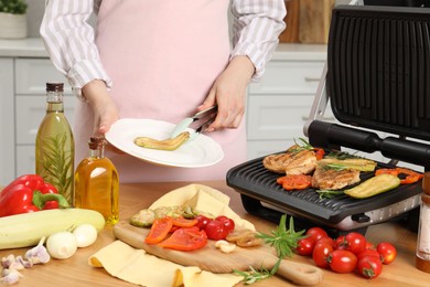 Woman cooking different products with electric grill at wooden table in kitchen, closeup