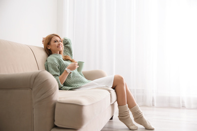 Young woman with cup of drink relaxing on couch near window at home. Space for text