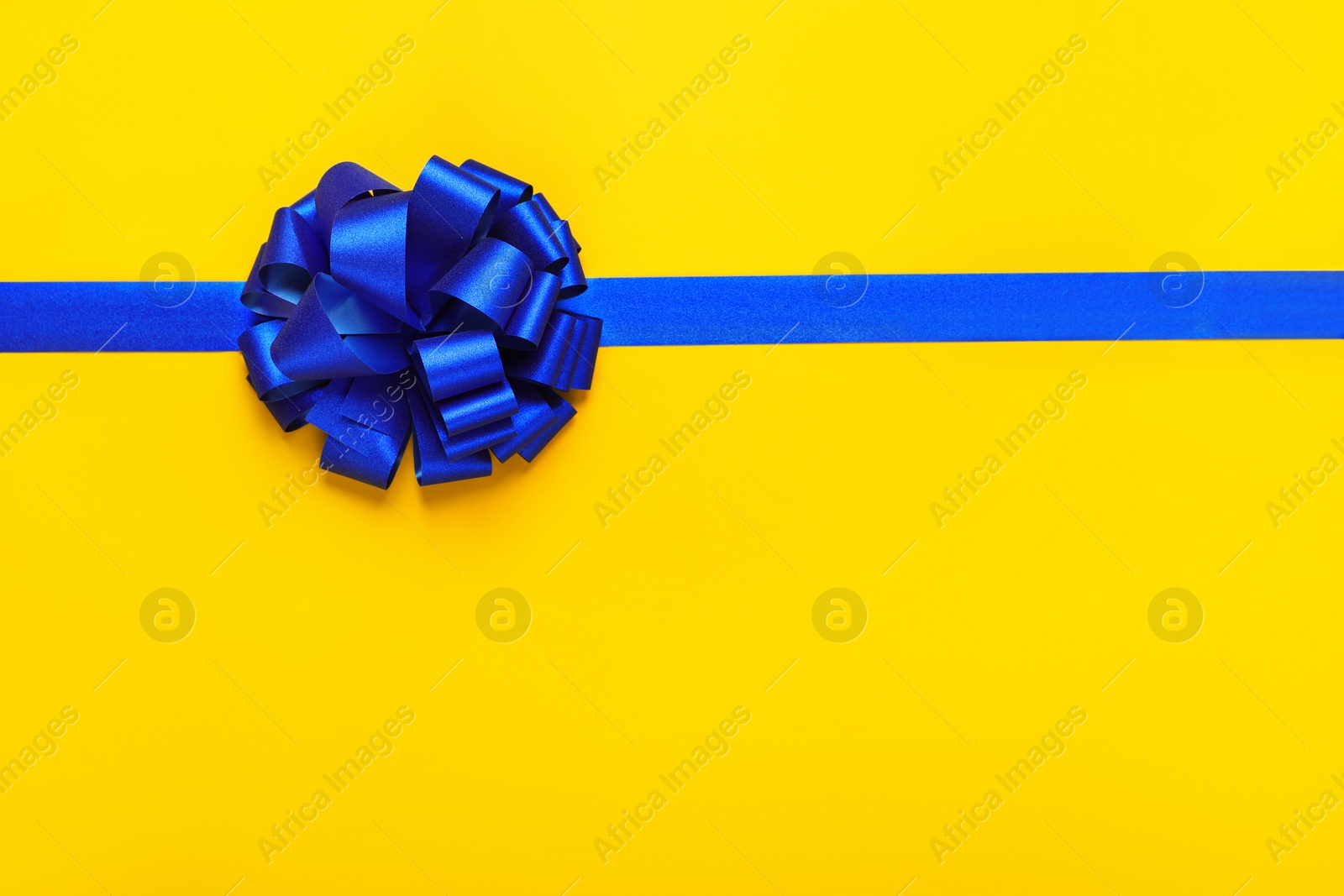 Photo of Blue ribbon with bow on yellow background, top view. Space for text