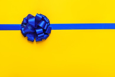 Photo of Blue ribbon with bow on yellow background, top view. Space for text