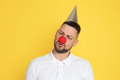 Photo of Funny man with clown nose and party hat on yellow background. April fool's day
