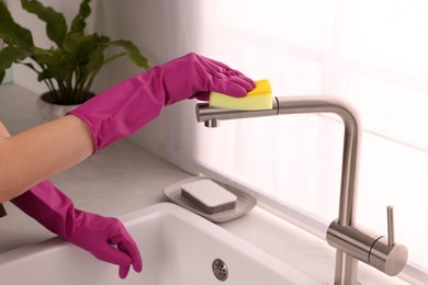 Photo of Woman in gloves cleaning faucet of kitchen sink with sponge, closeup