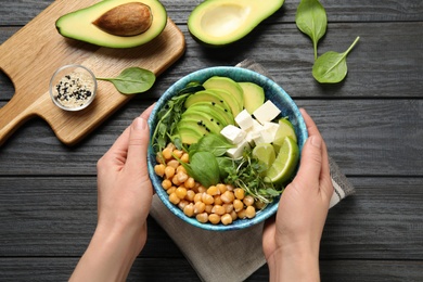 Photo of Woman holding bowl of delicious avocado salad with chickpea at black wooden table, top view