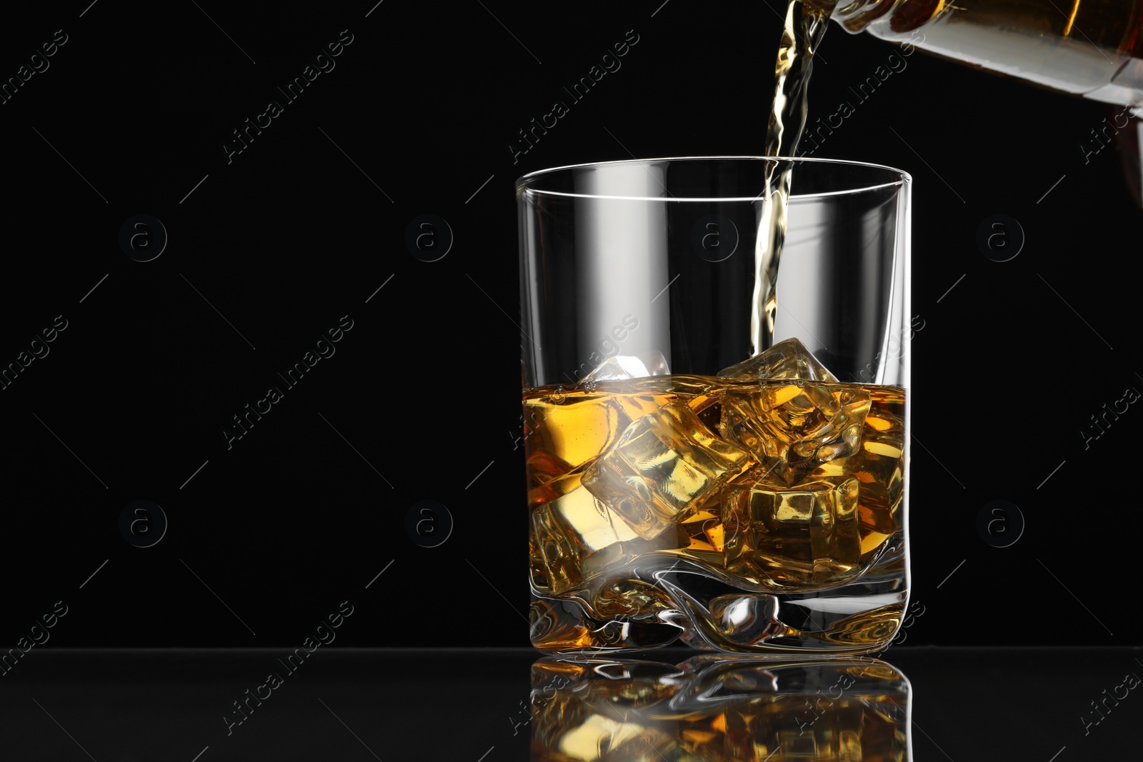 Photo of Pouring whiskey into glass with ice cubes at table against black background, space for text