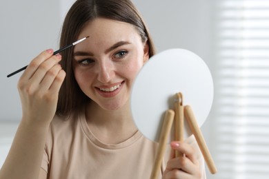 Photo of Smiling woman drawing freckles with brush in front of little mirror indoors