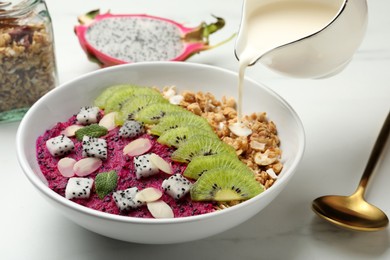 Photo of Adding milk to granola with pitahaya, kiwi and almonds on white table, closeup