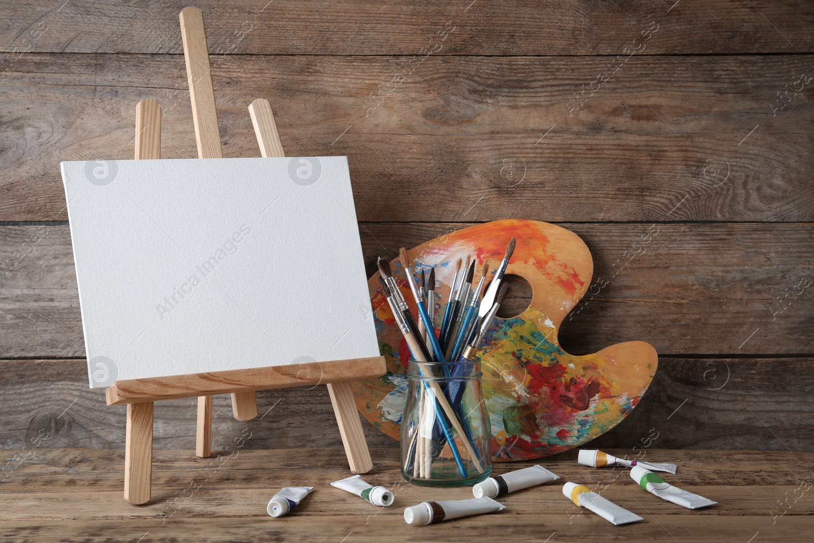 Photo of Easel with blank canvas, brushes, paints and palette on wooden table