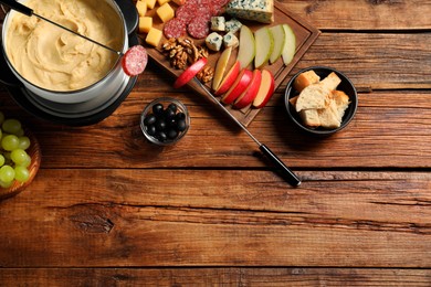 Fork with piece of sausage, melted cheese in fondue pot and other products on wooden table, flat lay. Space for text