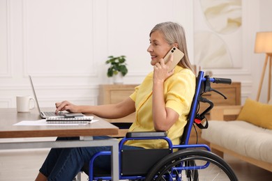 Woman in wheelchair talking on smartphone while using laptop at table in home office