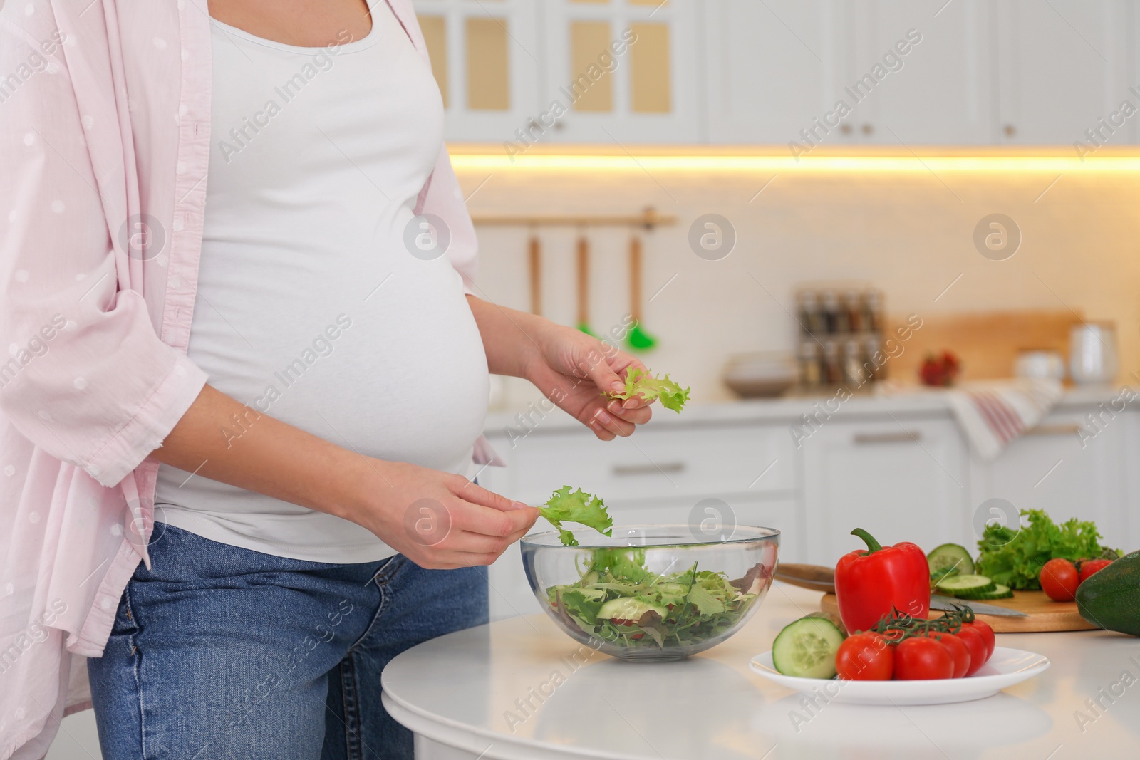 Photo of Young pregnant woman preparing vegetable salad at table in kitchen, closeup. Healthy eating