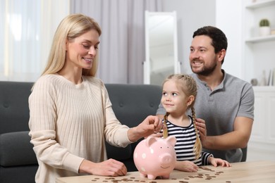 Photo of Family. Woman putting coin into piggy bank while her husband and daughter watching indoors