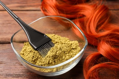 Photo of Bowl of henna powder, brush and red strand on wooden table, closeup. Natural hair coloring