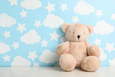 Photo of Teddy bear on white table near wall with painted blue sky, space for text. Baby room interior