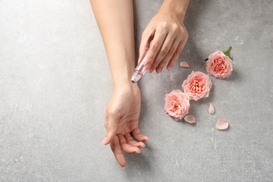 Photo of Woman applying rose essential oil on wrist and flowers at grey table, top view