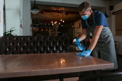 Photo of Waiter in mask and gloves disinfecting table at cafe