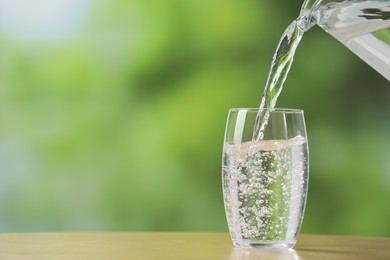 Pouring fresh water from jug into glass at wooden table against blurred green background, closeup. Space for text