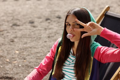 Photo of Beautiful young woman blowing chewing gum and showing peace gesture outdoors, space for text