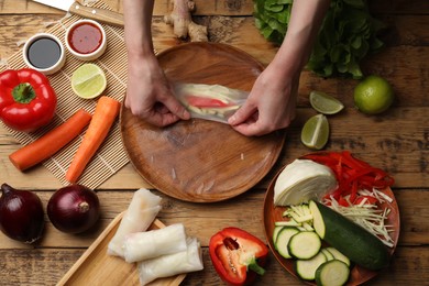 Photo of Making delicious spring rolls. Woman wrapping fresh vegetables into rice paper at wooden table, flat lay