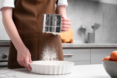 Woman sieving flour into baking dish at table in kitchen, closeup