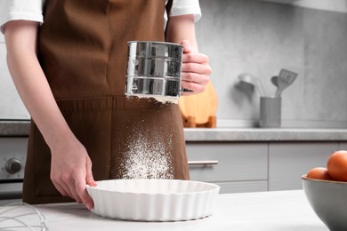 Photo of Woman sieving flour into baking dish at table in kitchen, closeup