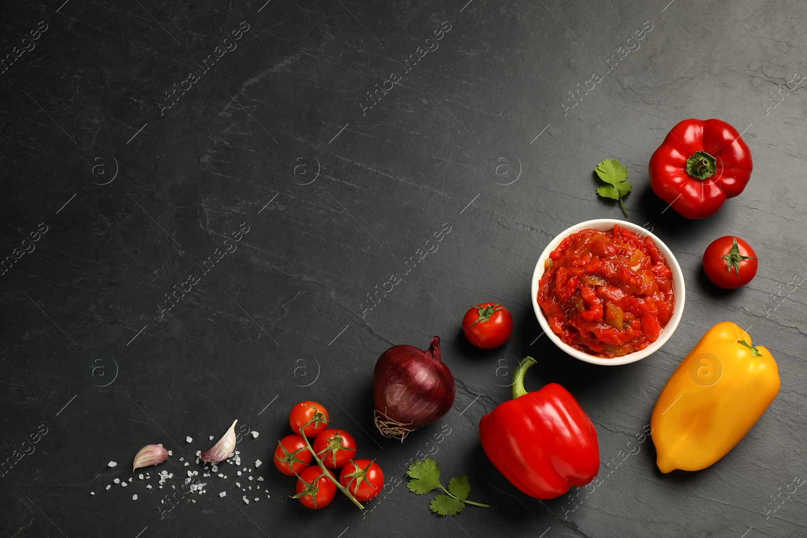 Photo of Delicious lecho in bowl and fresh ingredients on black table, flat lay. Space for text