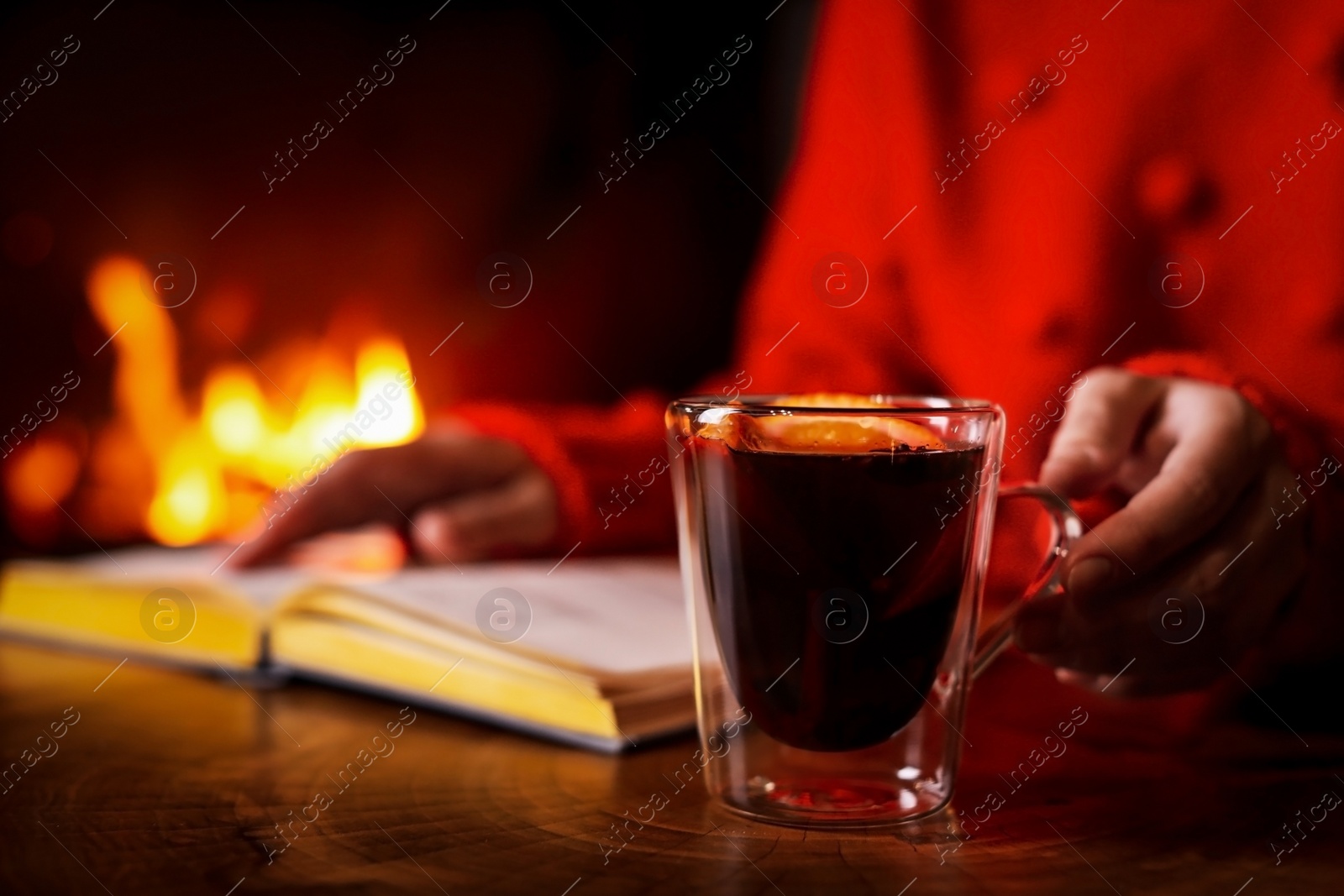 Photo of Woman with tasty mulled wine reading book near fireplace at home, closeup