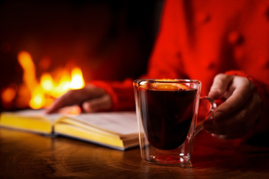 Photo of Woman with tasty mulled wine reading book near fireplace at home, closeup