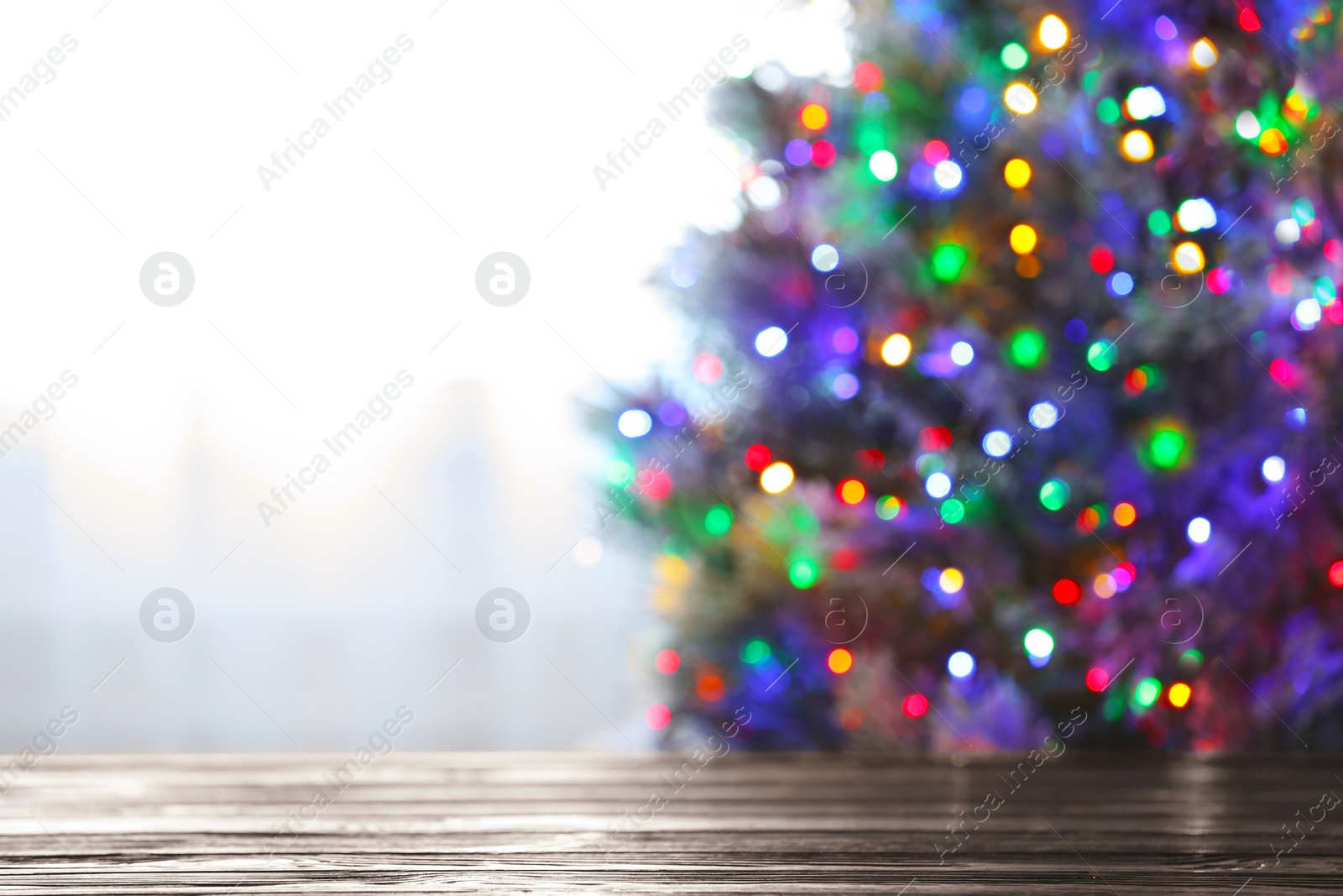 Photo of Blurred view of beautiful Christmas tree with colorful lights near window indoors, focus on wooden table. Space for text