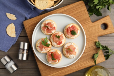 Photo of Delicious crackers with cream cheese, prosciutto and parsley on wooden table, flat lay