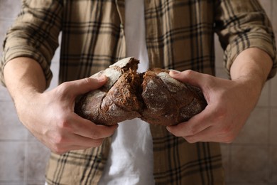 Photo of Man breaking loaf of fresh bread near grey wall, closeup