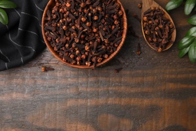 Aromatic cloves in bowl, spoon and green leaves on wooden table, flat lay. Space for text