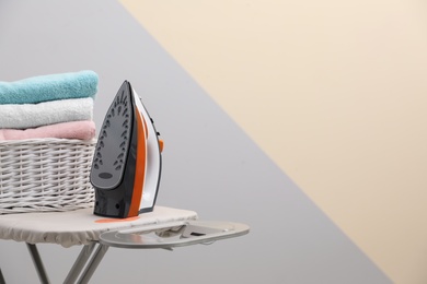 Photo of New modern iron and basket with laundry on board against color background, space for text