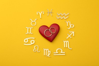 Zodiac signs, red heart and wedding rings on yellow background, flat lay