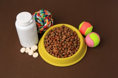 Photo of Bowl with dry pet food, bottle of vitamins and toys on brown background