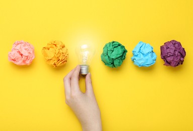 Photo of Woman holding lightbulb among colorful paper balls on yellow background, top view. Idea concept