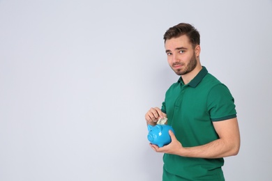 Young man putting money into piggy bank on light background. Space for text