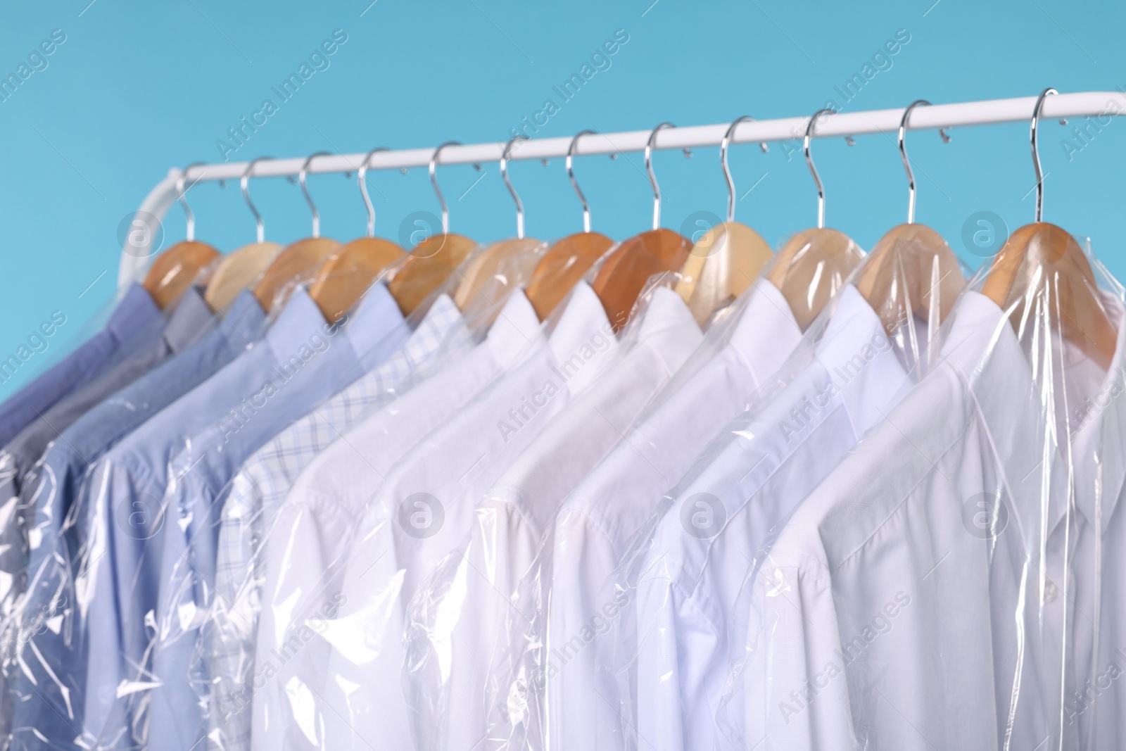 Photo of Dry-cleaning service. Many different clothes in plastic bags hanging on rack against light blue background