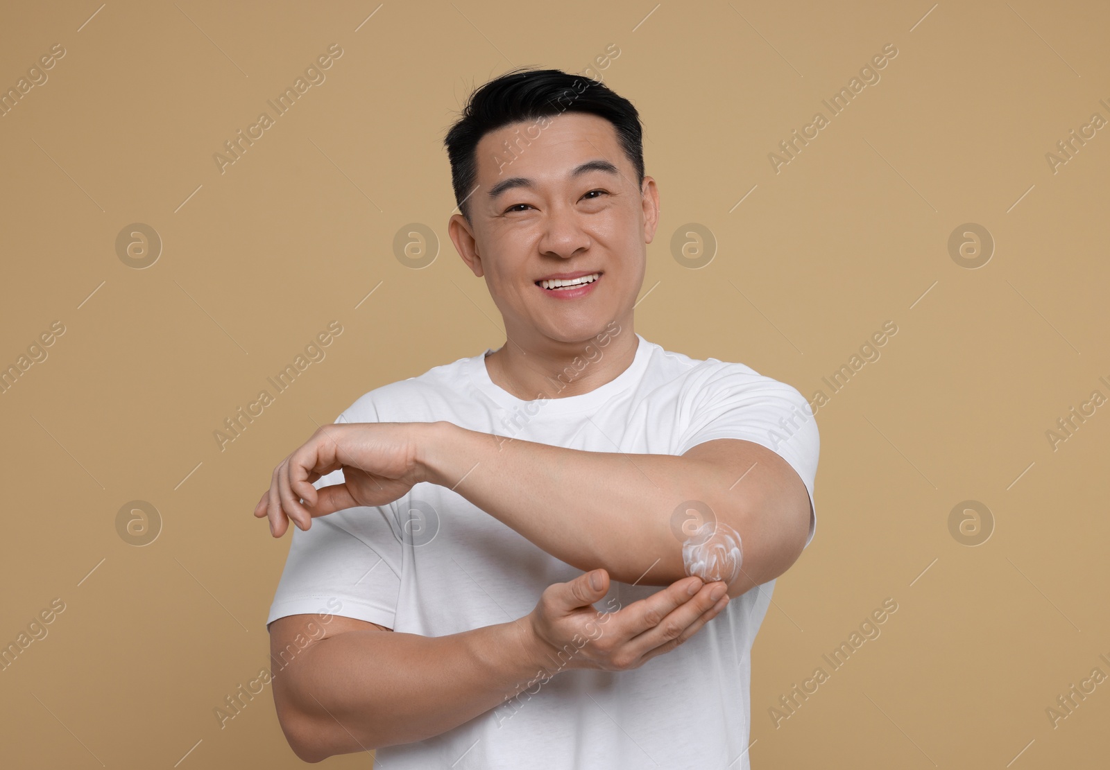 Photo of Handsome man applying body cream onto his elbow on light brown background