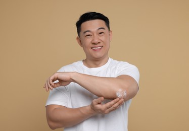 Photo of Handsome man applying body cream onto his elbow on light brown background