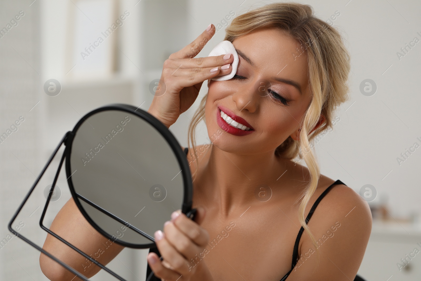 Photo of Smiling woman removing makeup with cotton pad in front of mirror indoors