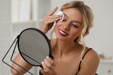 Smiling woman removing makeup with cotton pad in front of mirror indoors